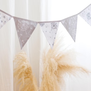 Könnte beinhalten: Ein weiß-graues Stoffbanner mit einem Löwenzahnmuster hängt vor einem weißen Hintergrund. Das Banner besteht aus Dreiecken und ist mit weißem Garn zusammengefügt.