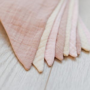 May include: A set of seven fabric pennants in various shades of pink and cream. The pennants are made of a soft, textured fabric and are arranged in a fan shape.