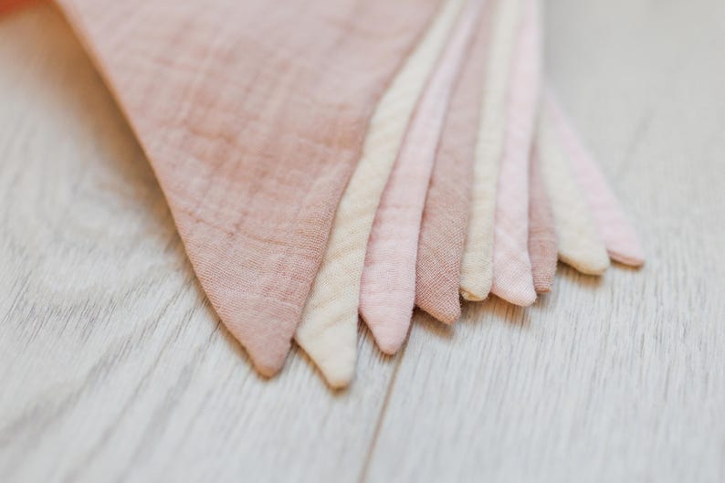 May include: A set of seven fabric pennants in various shades of pink and cream. The pennants are made of a soft, textured fabric and are arranged in a fan shape.