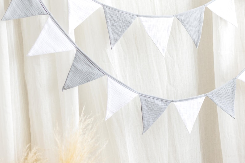 May include: A decorative banner with alternating white and gray triangular flags. The flags are strung together on a thin, light-colored string. The banner is set against a soft, white background.