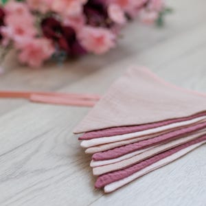 May include: A pink and mauve fabric bunting with a string attached. The bunting is made of multiple layers of fabric and is folded in half.
