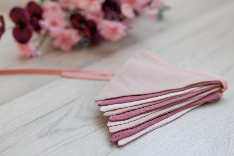 May include: A pink and mauve fabric bunting with a string attached. The bunting is made of multiple layers of fabric and is folded in half.