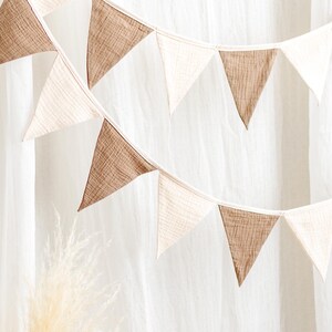 May include: Two strings of triangular fabric bunting in alternating shades of brown and off-white. The bunting is hanging in front of a white curtain. The fabric has a textured appearance.