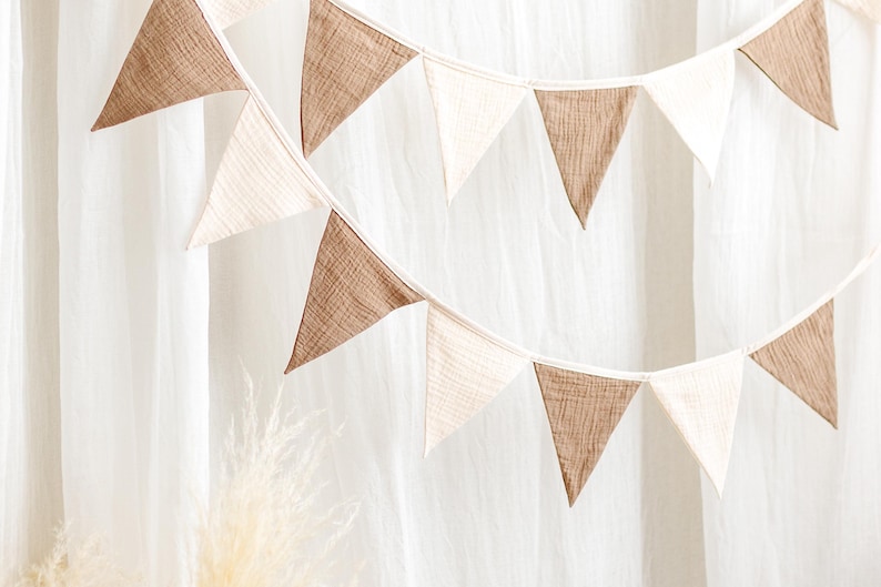 May include: Two strings of triangular fabric bunting in alternating shades of brown and off-white. The bunting is hanging in front of a white curtain. The fabric has a textured appearance.