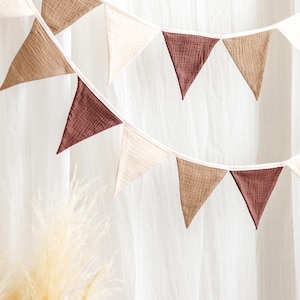 May include: A decorative banner with triangular flags in shades of brown, beige, and burgundy. The flags are strung together on a white string, creating a festive and stylish look. The banner is set against a white backdrop, with a hint of dried grass in the foreground.