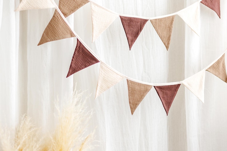 May include: A decorative banner with triangular flags in shades of brown, beige, and burgundy. The flags are strung together on a white string, creating a festive and stylish look. The banner is set against a white backdrop, with a hint of dried grass in the foreground.