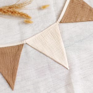May include: A neutral-toned fabric banner with alternating brown and cream-colored triangular flags. The banner is made of a textured material and is laid out on a white surface. Dried wheat stalks are placed near the banner.