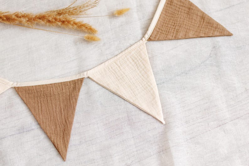 May include: A neutral-toned fabric banner with alternating brown and cream-colored triangular flags. The banner is made of a textured material and is laid out on a white surface. Dried wheat stalks are placed near the banner.