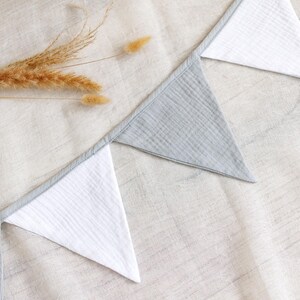 May include: A close-up shot of a decorative banner with alternating gray and white triangular flags. The banner is made of a textured fabric and is laid out on a neutral-colored surface. Dried wheat stalks are placed near the banner.