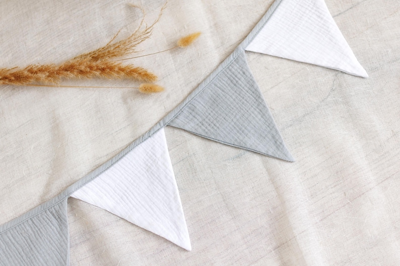 May include: A close-up shot of a decorative banner with alternating gray and white triangular flags. The banner is made of a textured fabric and is laid out on a neutral-colored surface. Dried wheat stalks are placed near the banner.