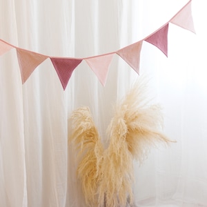 May include: A pink and white fabric bunting with triangular flags hanging against a white background. The bunting is made of soft, gauzy fabric and is perfect for adding a touch of whimsy to any room.