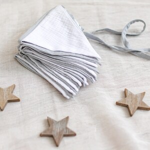 May include: A stack of white and gray muslin bibs with gray trim and ties. Three wooden star-shaped ornaments are scattered around the bibs on a neutral-colored surface. The bibs are folded neatly.
