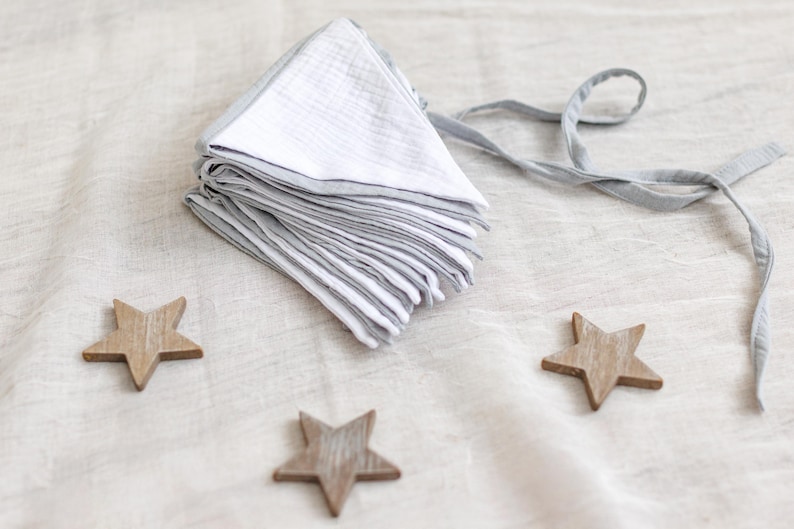 May include: A stack of white and gray muslin bibs with gray trim and ties. Three wooden star-shaped ornaments are scattered around the bibs on a neutral-colored surface. The bibs are folded neatly.