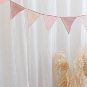 May include: A pink and white fabric bunting with triangle flags hanging against a white curtain backdrop. Two dried pampas grass stems are in the foreground.