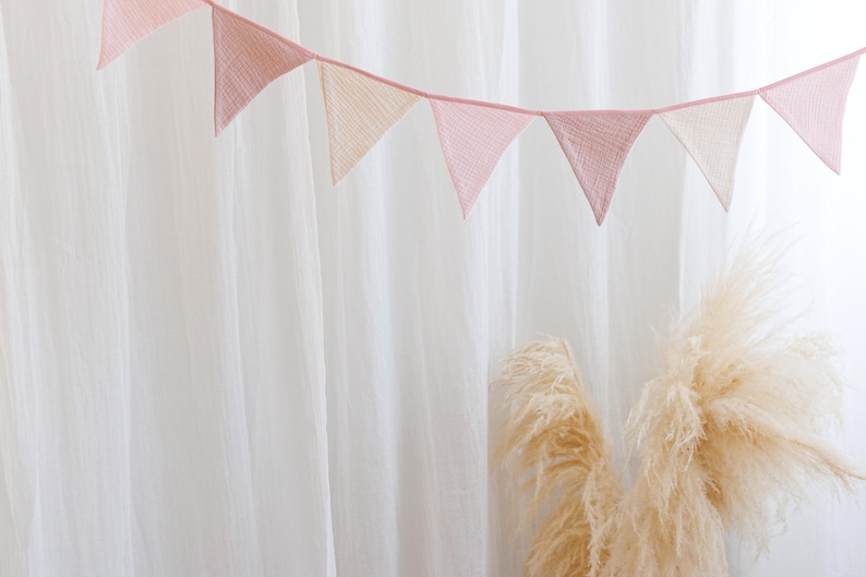May include: A pink and white fabric bunting with triangle flags hanging against a white curtain backdrop. Two dried pampas grass stems are in the foreground.
