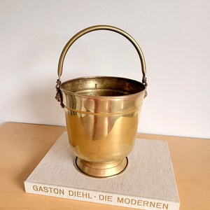 Op de afbeelding: Een vintage messing emmer met een gebogen handvat en decoratieve details. De emmer heeft een afgeronde basis en een gepolijste afwerking. De emmer staat op een boek met de tekst "GASTON DIEHL • DIE MODERNEN".
