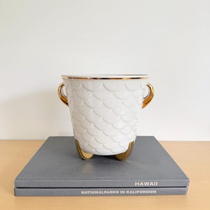 Pode incluir: Um recipiente de cerâmica branca com um padrão de escamas em relevo, apresentando alças douradas e três pés dourados. A borda é acentuada com uma fina faixa dourada. O recipiente tem um formato cônico e um acabamento liso e brilhante.