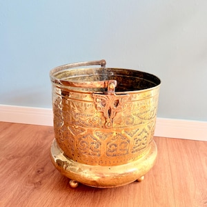 Peut inclure: Un seau en métal doré avec des motifs gravés complexes et une poignée. Le seau repose sur une base ronde avec de petits pieds. Le design présente des motifs floraux et géométriques, créant une esthétique vintage.