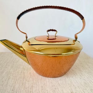Könnte beinhalten: Eine Vintage-Teekanne aus Kupfer und Messing mit geflochtenem Griff. Die Teekanne hat einen polierten Kupferkörper, einen Messingdeckel und einen Messingausguss. Der Griff besteht aus Kupfer und ist mit einem geflochtenen Material umwickelt.