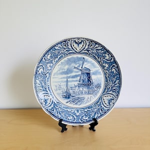Könnte beinhalten: Ein blau-weißes Zierteller mit einer Windmühlen-Szene. Der Teller hat einen detaillierten Rand mit floralen und scrollartigen Designs. Die Windmühle ist mit einem Segelboot und Gebäuden im Hintergrund dargestellt.