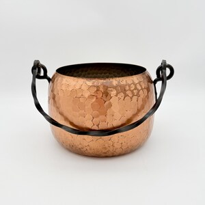 Puede incluir: Una olla redonda de cobre martillado con una abertura ancha y un asa de metal oscuro y retorcido sujeta a dos bucles laterales. La superficie exterior muestra una textura distintiva de hoyuelos sobre el acabado de cobre metálico brillante.