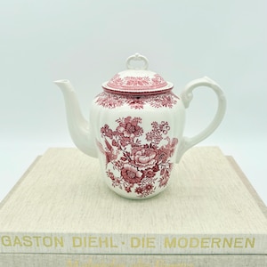 Könnte beinhalten: Weiße Keramik-Teekanne mit rotem Blumenmuster und Deckel. Die Teekanne hat einen gebogenen Griff und Ausguss. Das florale Design umfasst Vögel und Blumen. Die Teekanne steht auf einem Stapel Bücher.