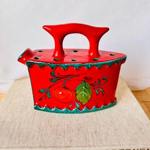 Könnte beinhalten: Rotes Keramikbügeleisen mit einem grünen und weißen Kirschendesign. Das Bügeleisen hat einen Griff und Löcher oben für Blumenarrangements.