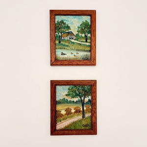 Puede incluir: Dos pinturas de paisajes enmarcadas. La pintura superior representa una casa con tejado de paja, árboles y patos en un estanque. La pintura inferior muestra balas de heno, un camino y un árbol. Ambas están en marcos de madera marrón.