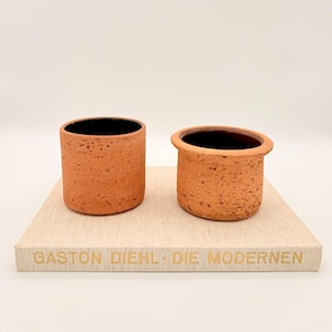 Op de afbeelding: Twee terracotta keramische potten met zwarte binnenkanten. Eén is cilindrisch, de andere heeft een afgeronde rand. Ze staan op een lichtgekleurde rechthoekige basis met gouden tekst "GASTON DIEHL - DIE MODERNEN".