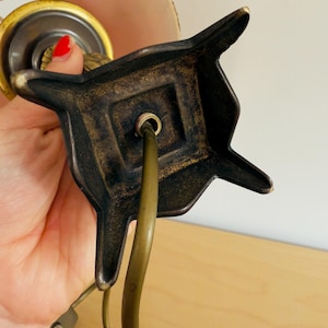 Pu&ograve; includere: Primo piano di una base di lampada vintage con finitura bronzo scuro. La base ha un design decorativo a forma di stella con un quadrato centrale. Un cavo e un interruttore color ottone sono visibili, suggerendo un apparecchio di illuminazione antico.