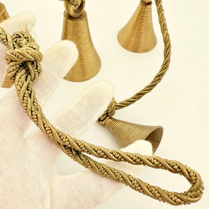 May include: Close-up of several small, gold-colored bells with a twisted rope design. The bells have a conical shape and are suspended from a knotted rope. The background is a plain white surface.