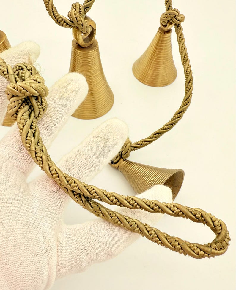 May include: Close-up of several small, gold-colored bells with a twisted rope design. The bells have a conical shape and are suspended from a knotted rope. The background is a plain white surface.