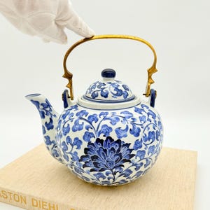 Op de afbeelding: Een blauw-witte porseleinen theepot met een bloemmotief en een goudkleurige handgreep. De theepot heeft een ronde vorm en een deksel met een kleine blauwe knop. Het bloemmotief is in blauwtinten op een witte achtergrond.