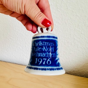 Może przedstawiać: Biały porcelanowy dzwonek z ciemnoniebieskim ozdobnym pasem zawierającym napis Christmas, Jule, Noel, Weihnachten oraz rok 1976. Dzwonek posiada małą pętelkę na górze do zawieszenia oraz rozszerzaną podstawę z powtarzającym się niebieskim wzorem.