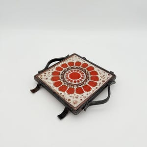Puede incluir: Salvamanteles cuadrado con un diseño decorativo de azulejo floral rojo y blanco. El azulejo está colocado en un marco de metal oscuro con asas y patas curvas. Un accesorio de cocina de estilo vintage.