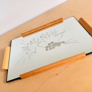 Op de afbeelding: Een rechthoekige spiegellade met houten handvatten. De spiegel heeft een geëtst ontwerp van sterren en cirkels. De lichtbruine houten handvatten zijn aan de zijkanten van de spiegel bevestigd.