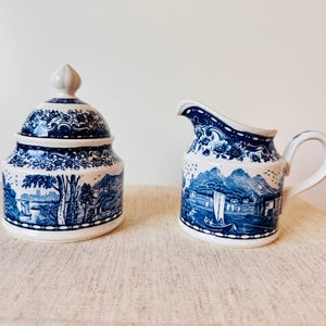 Puede incluir: Un juego de azucarero con tapa y jarra para crema a juego. Ambas piezas son blancas con un diseño de paisaje azul. El azucarero tiene forma redondeada con tapa y un pequeño adorno. La jarra para crema tiene asa y pico.