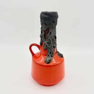 May include: A ceramic jug with a vibrant orange base and a single handle. The neck of the jug features a thick, dark grey, porous lava-like glaze that drips down onto the smooth, glossy orange body, creating a distinct textural contrast.