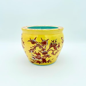 Pode incluir: Um vaso de cerâmica amarelo com um design floral em tons de vermelho, castanho e branco. O vaso tem um interior turquesa e uma borda decorativa. O vaso tem uma forma redonda.