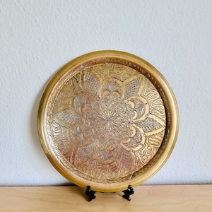 May include: A circular, gold-toned metal tray featuring an elaborate, etched floral pattern. The tray has a raised rim and is presented on a black stand. The surface of the tray displays a detailed, symmetrical design.
