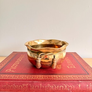 May include: A small, gold-colored bowl with a ruffled rim and decorative rope and tassel accents. The bowl is set atop a stack of vintage books with red covers and gold lettering. The books add a touch of antique charm.