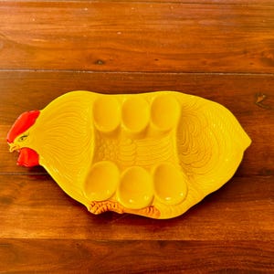 May include: A yellow ceramic egg holder in the shape of a rooster. The holder has six egg-shaped indentations and a red comb and wattle. The rooster-shaped holder is on a wooden surface.