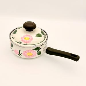 Può includere: Una casseruola in smalto bianco con coperchio, con un design floreale con fiori rosa e foglie verdi. Il manico è marrone scuro. La casseruola è progettata per cucinare.