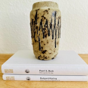 Puede incluir: Un jarrón de cerámica beige con detalles de esmalte negro goteado. El jarrón es cilíndrico con una parte superior ligeramente ensanchada. Se encuentra sobre dos libros blancos con los títulos "Pearl S. Buck Die gute Erde" y "Rudyard Kipling Das Dschungelbuch".