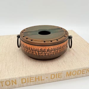 Könnte beinhalten: Ein kupferfarbener, runder Behälter mit Deckel. Der Deckel hat eine zentrale Öffnung und eine grüne und kupferfarbene Oberfläche. Die Seite des Behälters weist ein detailliertes, geprägtes Design auf. Zwei schwarze Griffe sind angebracht. Der Behälter steht auf einer hellen Oberfläche.