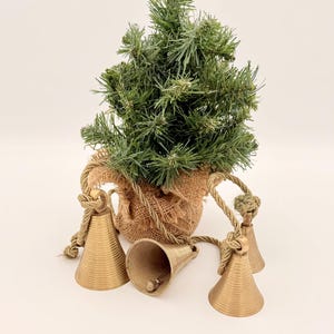 May include: A small artificial Christmas tree in a burlap sack with three gold-colored bells. The bells are attached to the sack with rope. The tree is green and has a natural look.