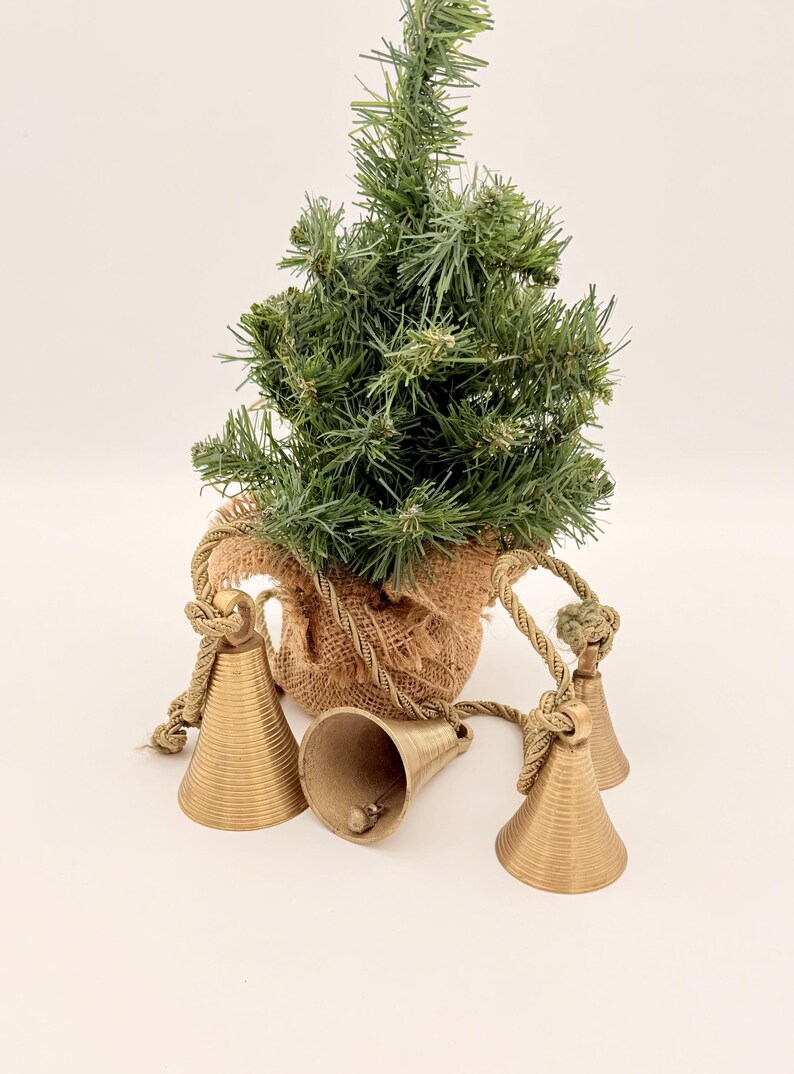 May include: A small artificial Christmas tree in a burlap sack with three gold-colored bells. The bells are attached to the sack with rope. The tree is green and has a natural look.