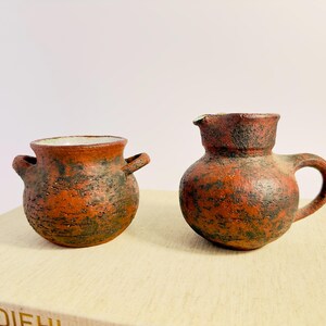 Peut inclure: Deux récipients en céramique rustiques faits à la main. L'un est un petit pot avec deux anses, l'autre une cruche avec une anse et un bec verseur. Les deux ont une surface texturée brun-rougeâtre avec des accents vert foncé.
