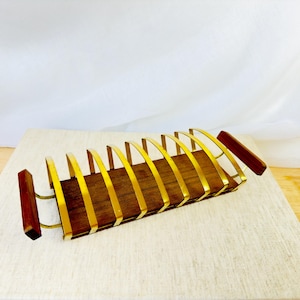 May include: A rectangular toast rack featuring a dark brown wooden base and two matching wooden handles on the ends. The structure consists of eight curved, gold-toned metal dividers that create slots for holding slices of toast upright.
