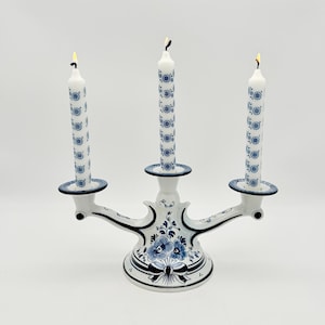 Peut inclure: Un candélabre en céramique blanche à trois branches orné de motifs floraux bleus et d'un nœud à la base. Trois bougies effilées blanches avec un imprimé floral bleu répétitif sont insérées dans les supports, chacune surmontée d'une flamme.